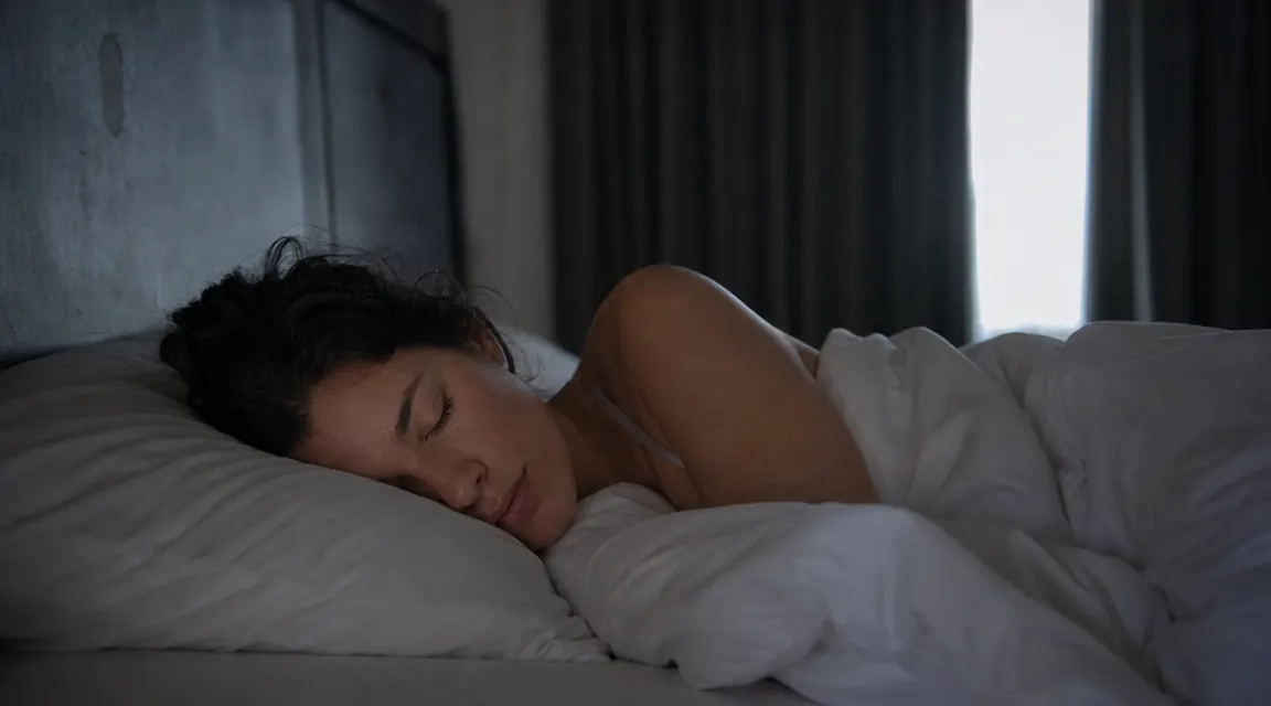 Person sleeping peacefully in a dark bedroom with soft morning light beginning to show at the curtain edges