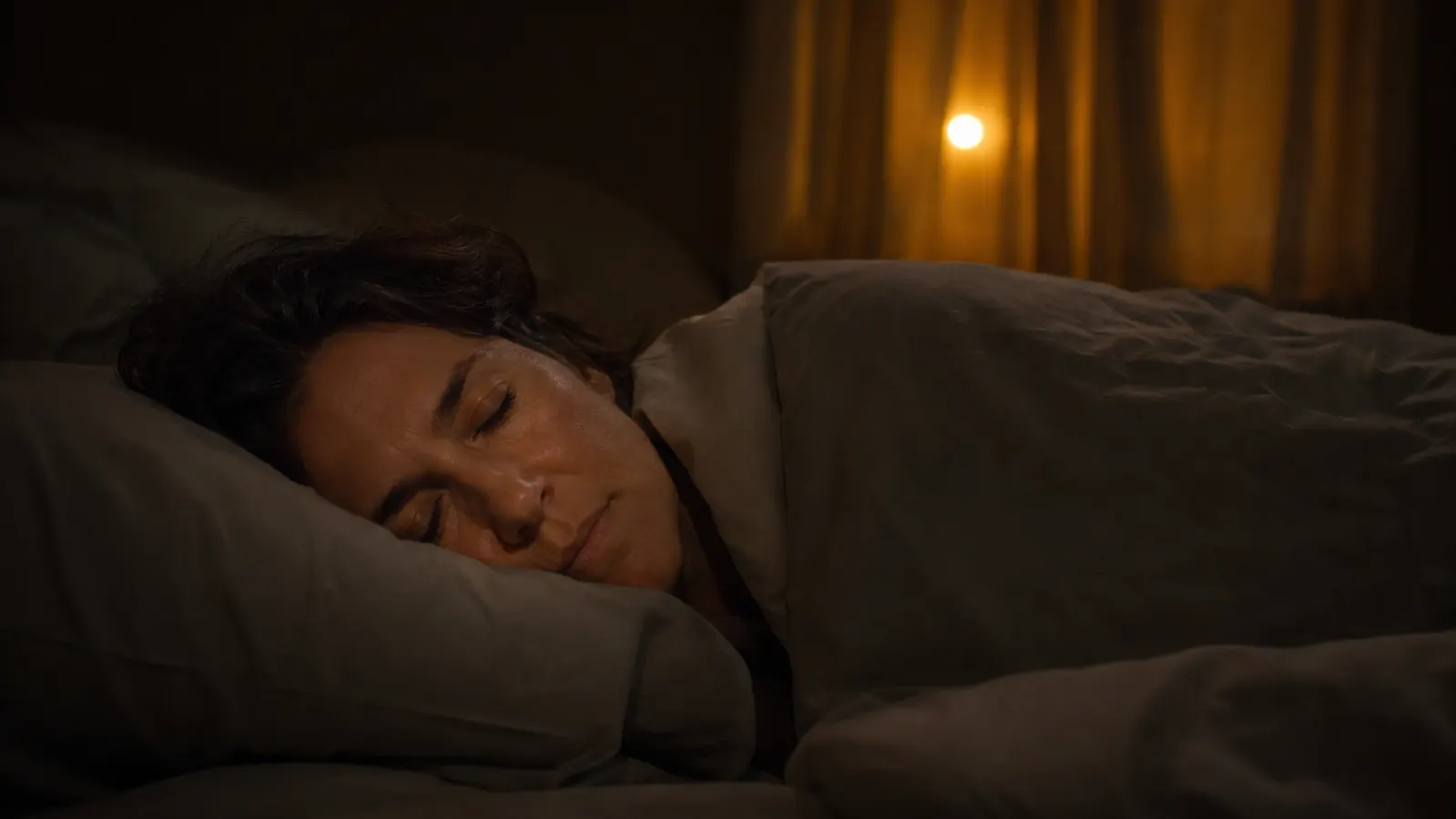 Woman sleeping peacefully in a dark bedroom, soft predawn light visible through curtains behind her